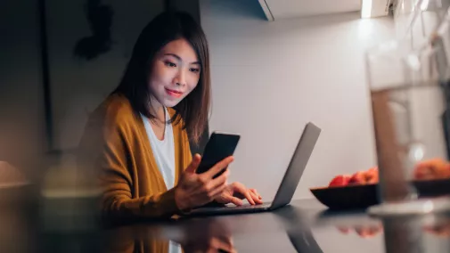 Frau schaut auf Smartphone und sitzt mit Computer zu Hause 