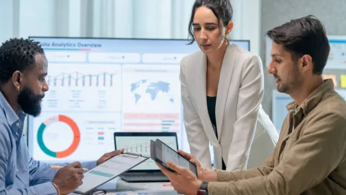 Three people are sitting in a meeting room looking at charts on a tablet and laptop.