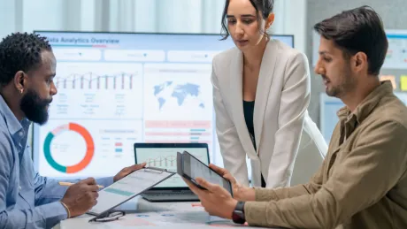 Drei Personen sitzen im Meetingraum und betrachten Diagramme auf einem Tablet und Laptop.