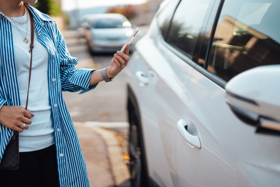 A woman is standing in front of her car, holding her smartphone in her hand.